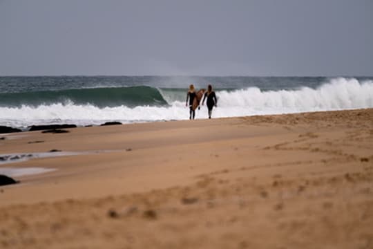 Two surfers walking along a sandy beach with waves breaking offshore