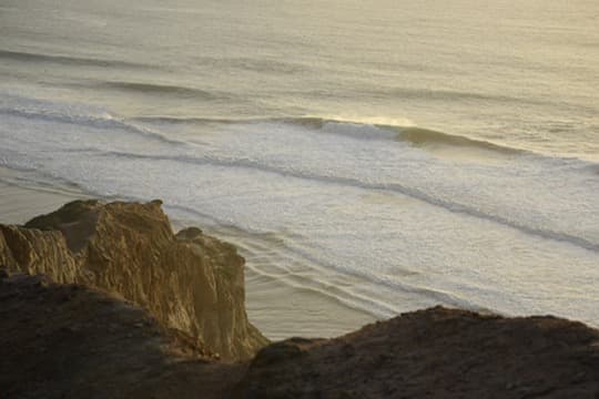 Long lines of swell wrapping toward a beach below coastal cliffs
