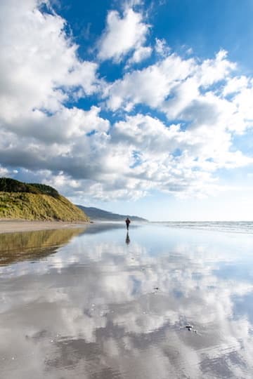 Person walking across a reflective beach beneath a bright sky