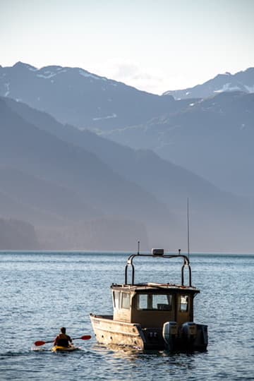 Small skiff and kayaker on calm water beneath coastal mountains