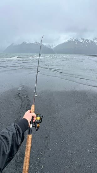 Angler casting from a dark beach toward snowy coastal mountains