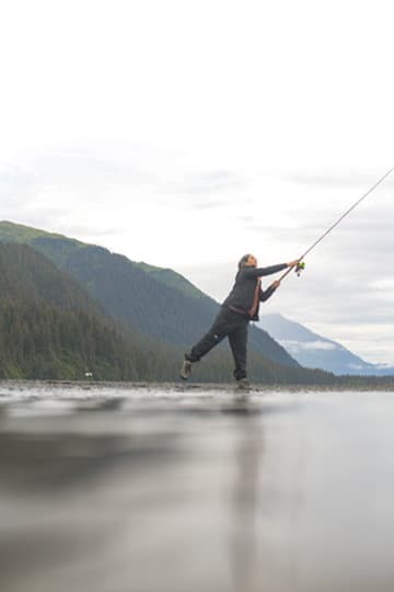 Angler casting a fly rod in shallow water beside a forested coast